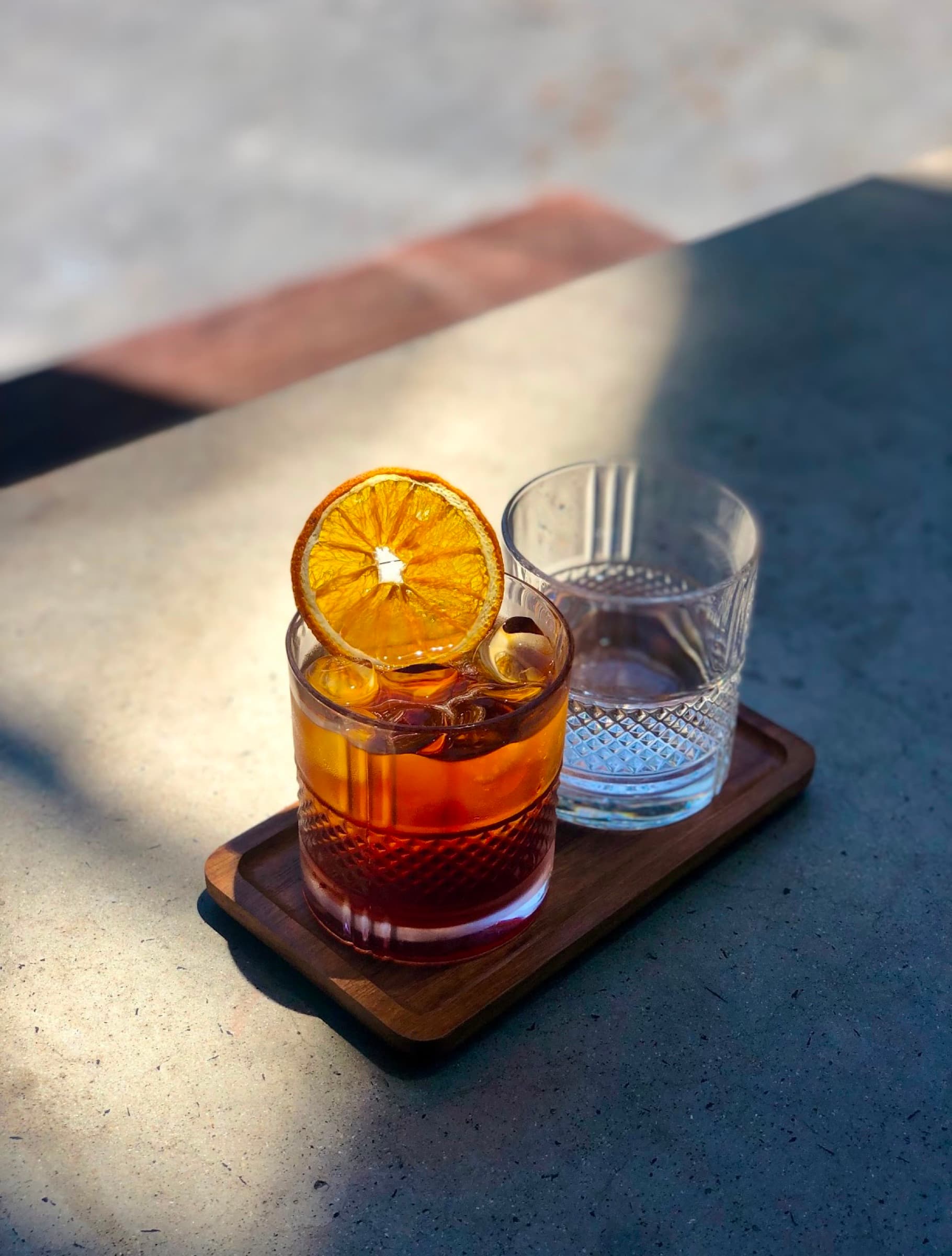 A Negroni with a dried orange wheel on a wooden serving board, water carafe beside it, sunlight cutting across