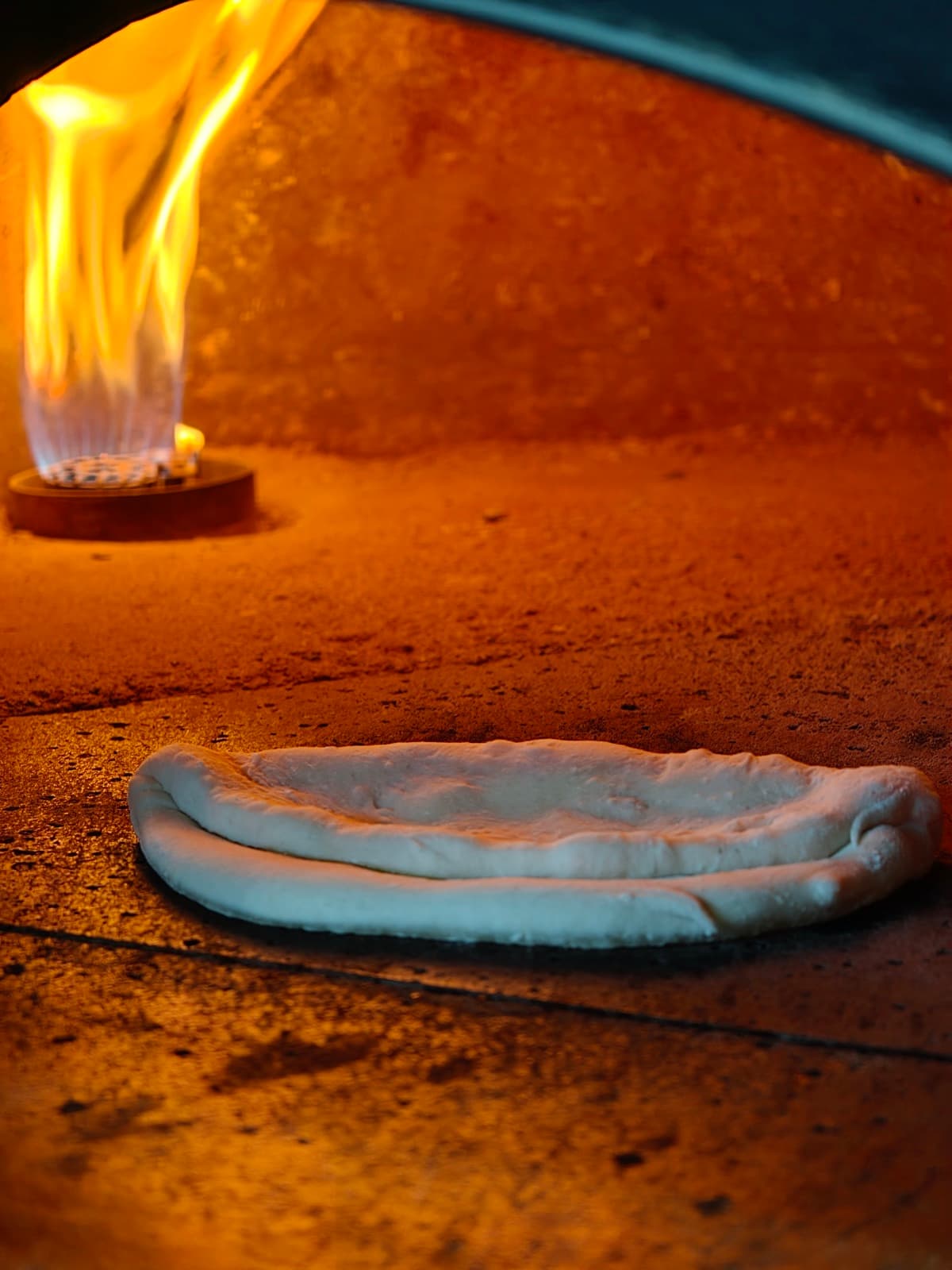 An Ozzo base baking on the floor of the wood-fired oven, flames rising against the dome behind it
